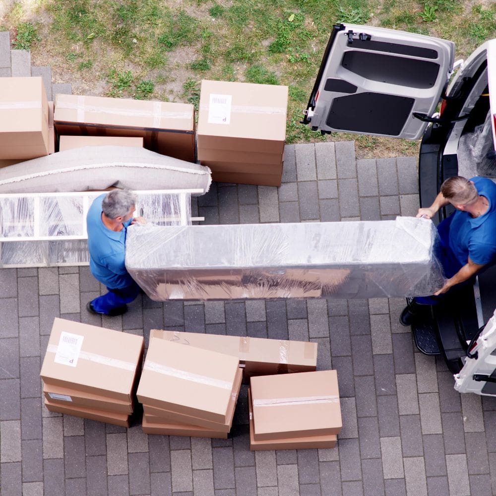 Dos operarios de mudanzas cargando un sofá protegido con film plástico en una furgoneta blanca en un chalet de La Moraleja, Madrid.