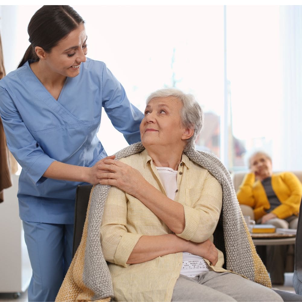 Una cuidadora con uniforme azul sonríe mientras coloca una manta sobre los hombros de una mujer mayor en un salón iluminado en La Moraleja.