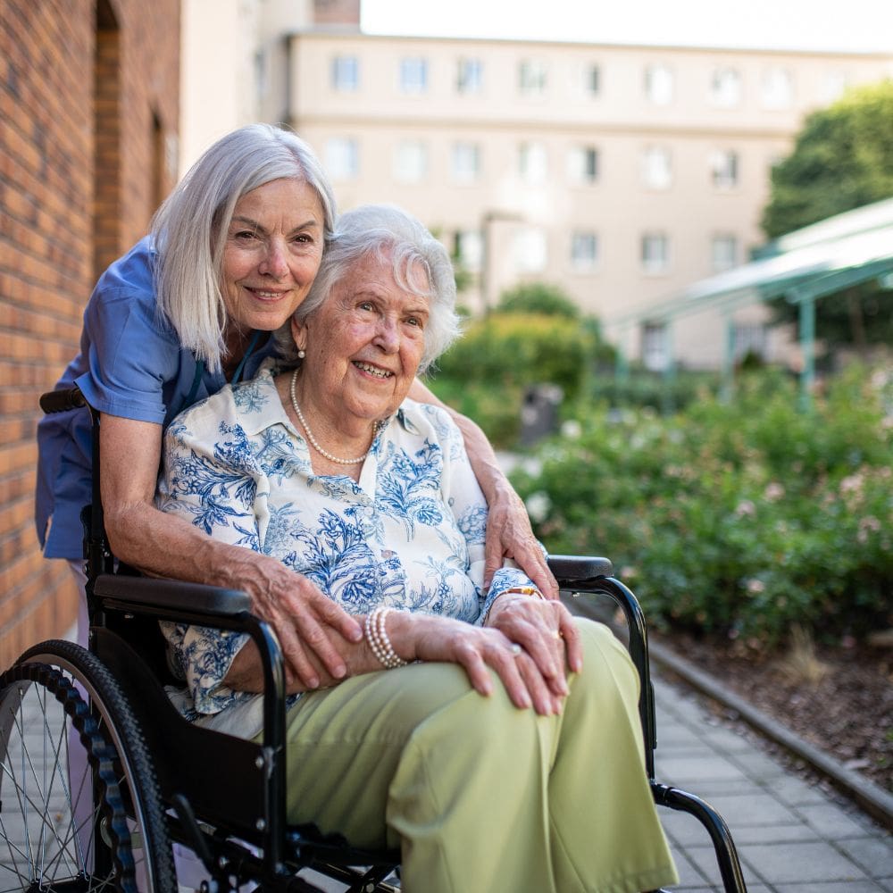 Cuidadora con uniforme azul sonriendo junto a mujer mayor en silla de ruedas en un jardín de La Moraleja.