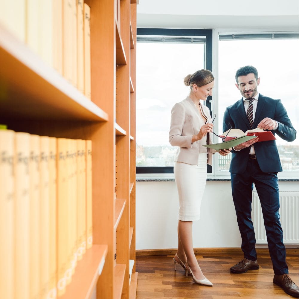 Un abogado y una abogada de un bufete en La Moraleja consultando documentación legal frente a una estantería de libros jurídicos en un despacho moderno.